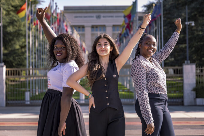 Drie meisjes staan voor het gebouw van de Verenigde Naties in Genève. Ze steken alledrie een hand in de lucht.